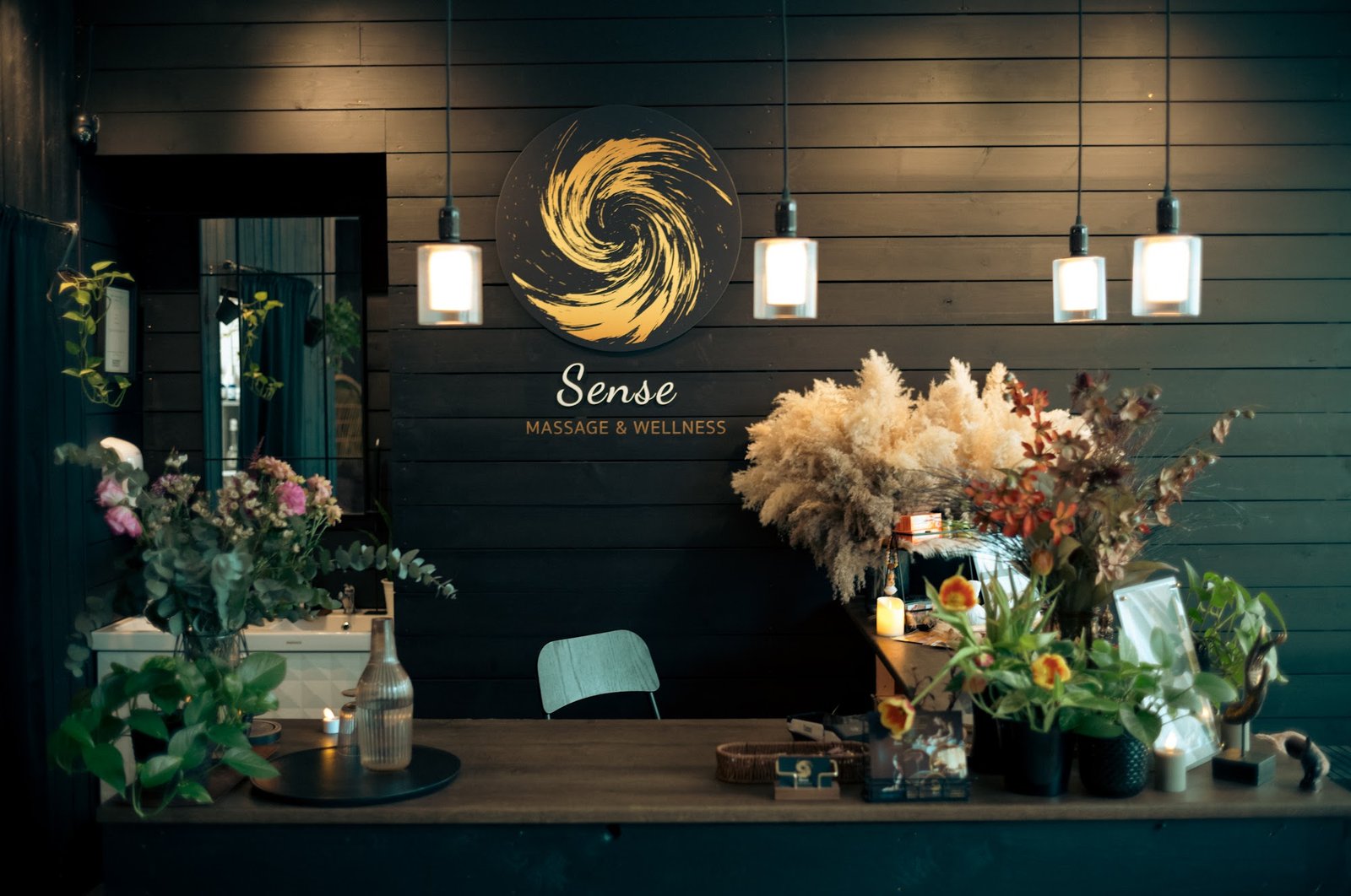 The Sense reception desk: dark wood-paneled wall with the gold Sense logo, pendant lights, dried pampas and fresh flowers